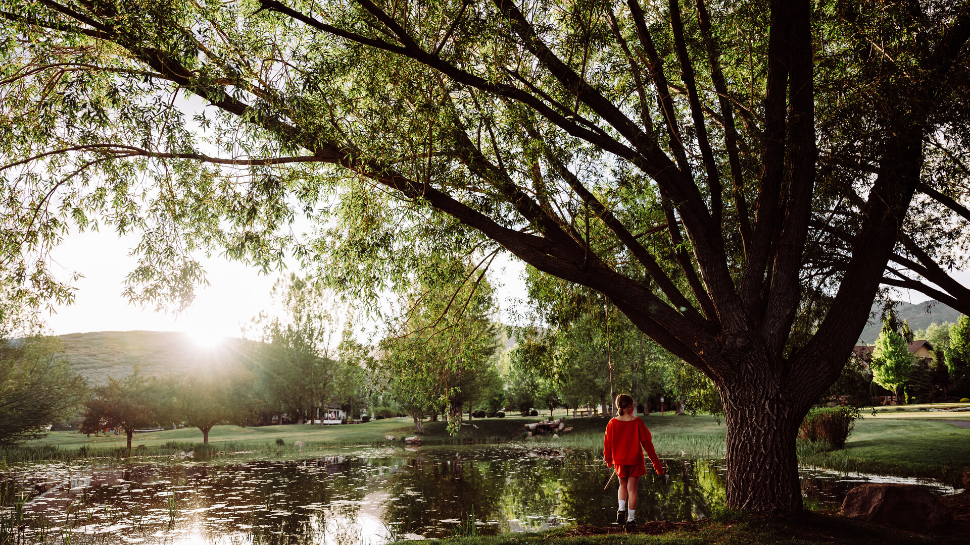 A child stands at the edge of a neighborhood pond in the Mountain West at golden hour, fishing pole in hand, willow tree arching overhead with a rope swing, Wasatch foothills on the horizon — a summer evening worth protecting.